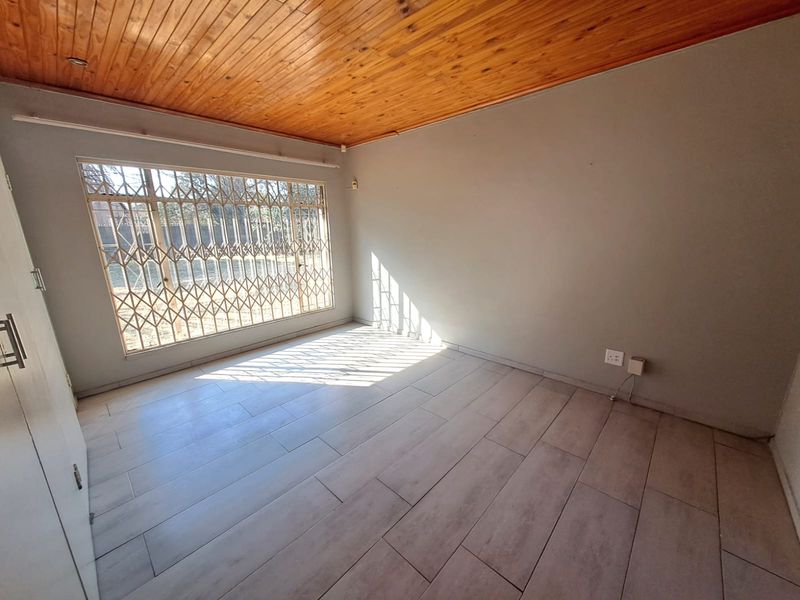A bright, empty bedroom featuring tiled flooring, a timber tongue and groove ceiling, and a large window fitted with a security gate.