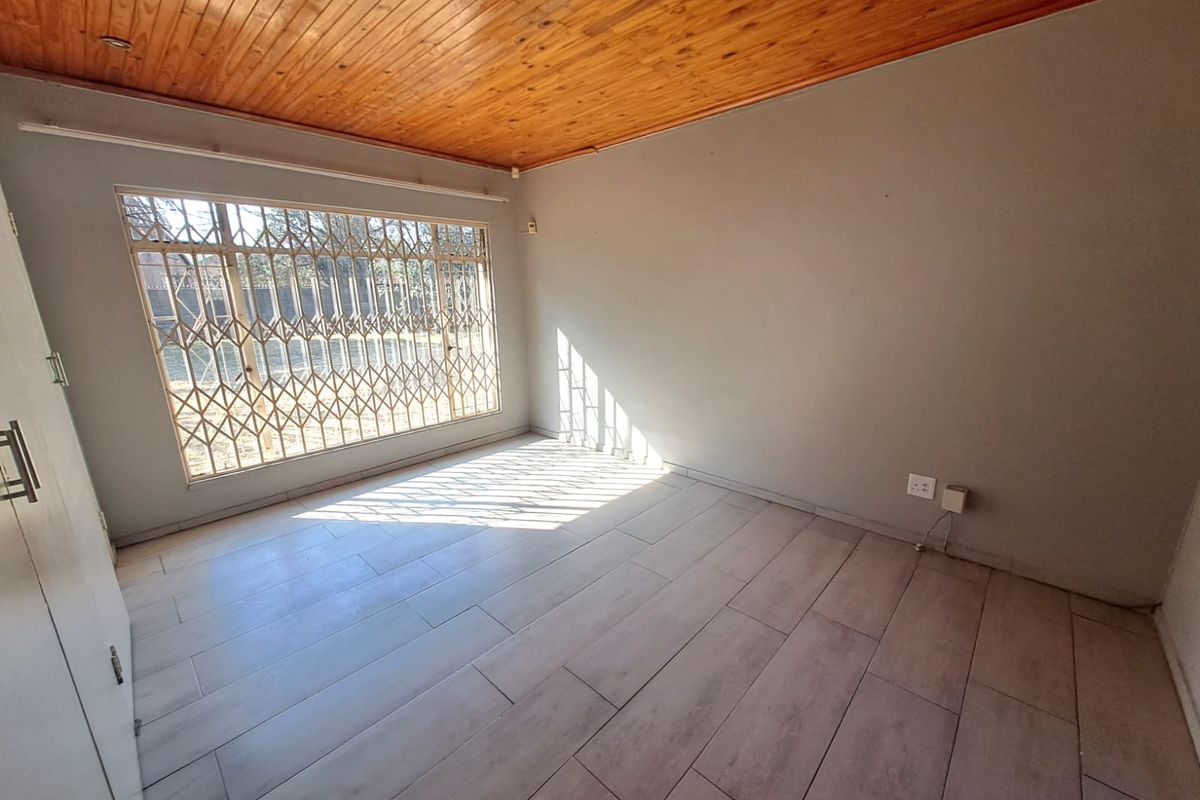 A bright, empty bedroom featuring tiled flooring, a timber tongue and groove ceiling, and a large window fitted with a security gate.