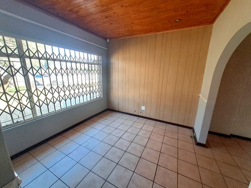 An empty living room featuring tiled flooring, a timber ceiling, and wood paneling, with a security gate on the window and an arched entryway.