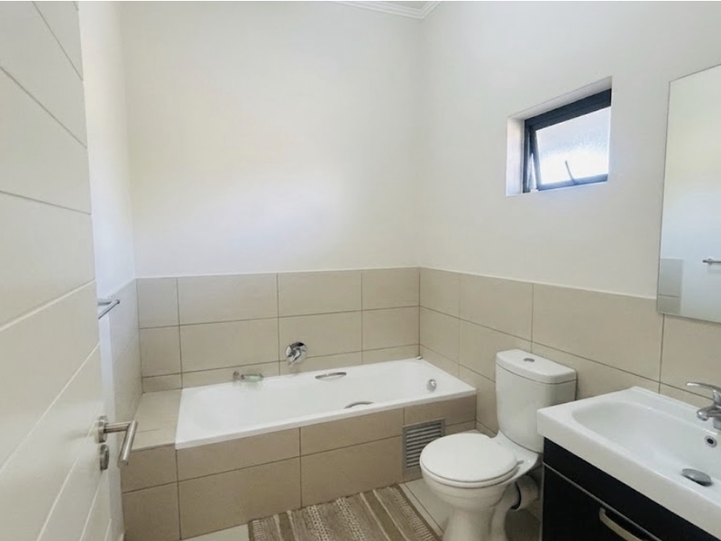 A clean, modern bathroom featuring a built-in bathtub, vanity basin with storage cabinet, and neutral wall tiling.