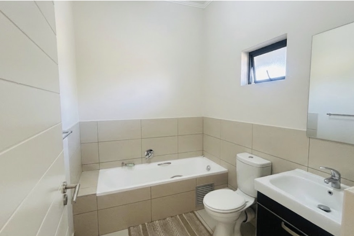 A clean, modern bathroom featuring a built-in bathtub, vanity basin with storage cabinet, and neutral wall tiling.