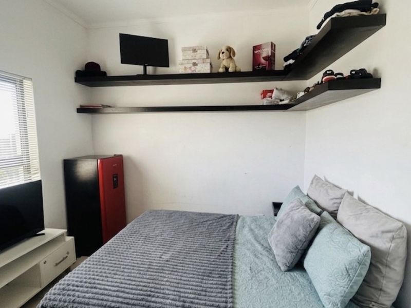 A compact bedroom featuring white walls, floating corner shelving, and a window with fitted blinds.