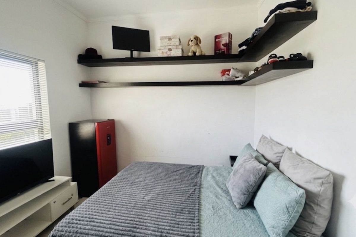 A compact bedroom featuring white walls, floating corner shelving, and a window with fitted blinds.