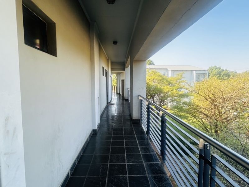 A covered outdoor balcony featuring dark tiled flooring and a metal balustrade, providing an open walkway with views of the surrounding greenery.