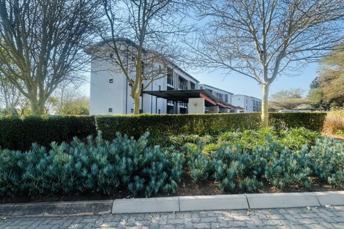 A view of the front elevation of a multi-story flat complex, featuring well-maintained landscaped gardens and paved driveway access.
