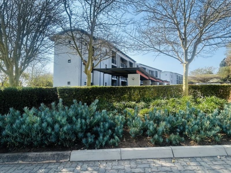 A view of the front elevation of a multi-story flat complex, featuring well-maintained landscaped gardens and paved driveway access.