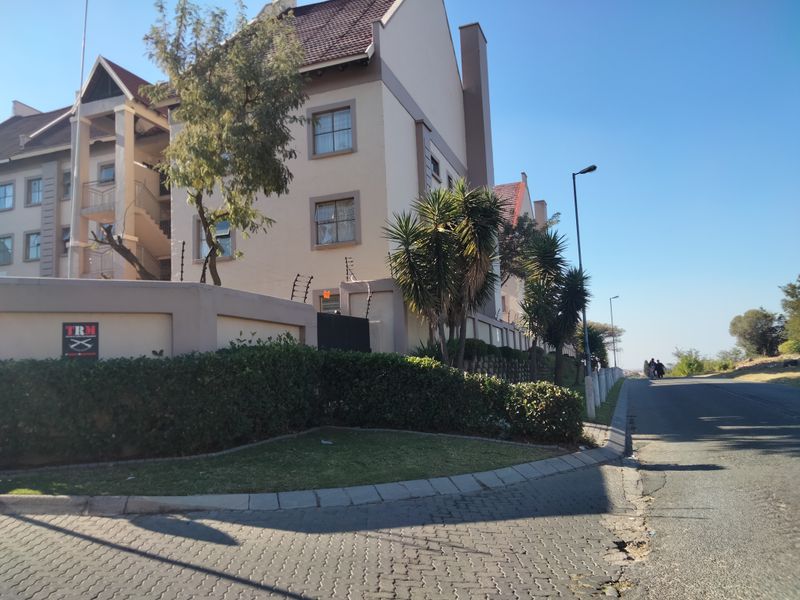 Exterior view of the apartment complex featuring a perimeter wall with electric fencing, paved driveway, and landscaped garden area.