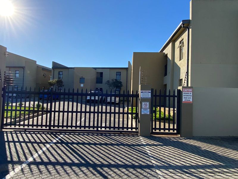 Front entrance of the apartment complex featuring an automated security gate, electric fencing, and a paved driveway.