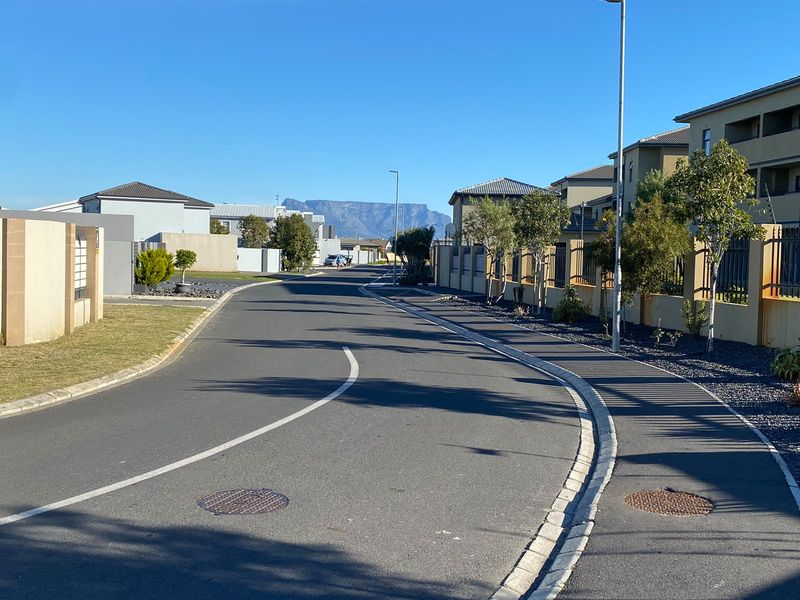 A clear street view of the residential complex featuring paved roads, perimeter walls, and mountain views in the distance.