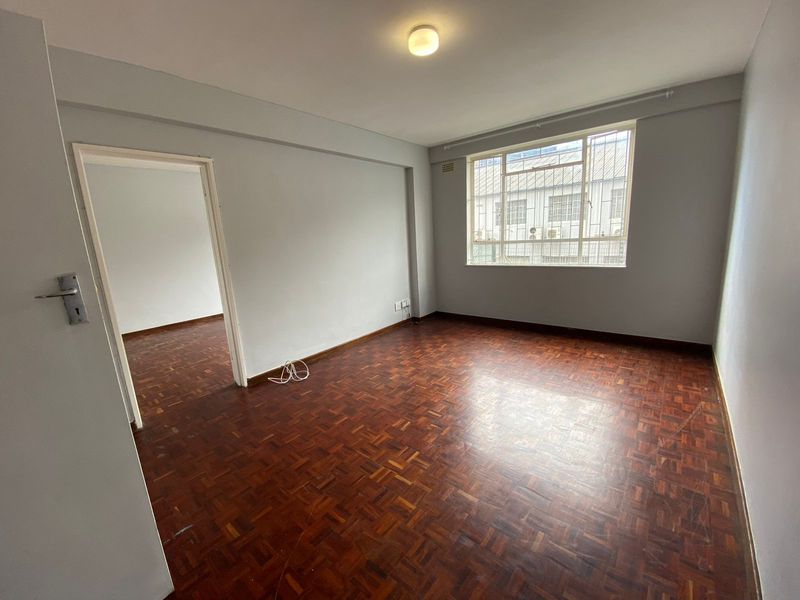 An empty living room featuring original parquet flooring, neutral wall paint, and a window fitted with security bars.