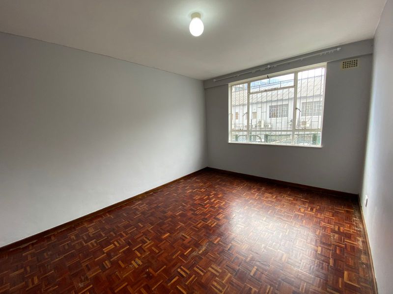 An empty bedroom featuring traditional parquet flooring, a large window with security bars, and neutral wall finishes.