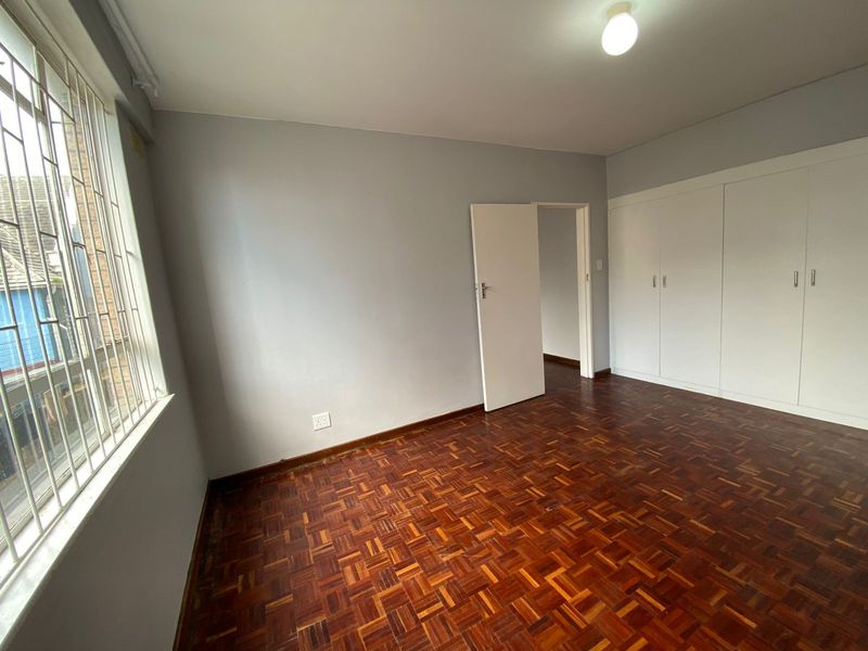 A bright, empty bedroom featuring classic parquet flooring, built-in storage cupboards, and a window fitted with security bars.