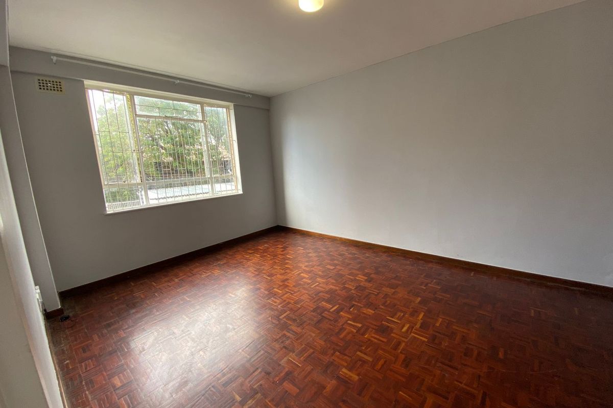 An empty bedroom featuring classic parquet flooring, a large window with security bars, and neutral wall finishes.
