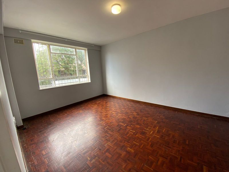 An empty bedroom featuring classic parquet flooring, a large window with security bars, and neutral wall finishes.