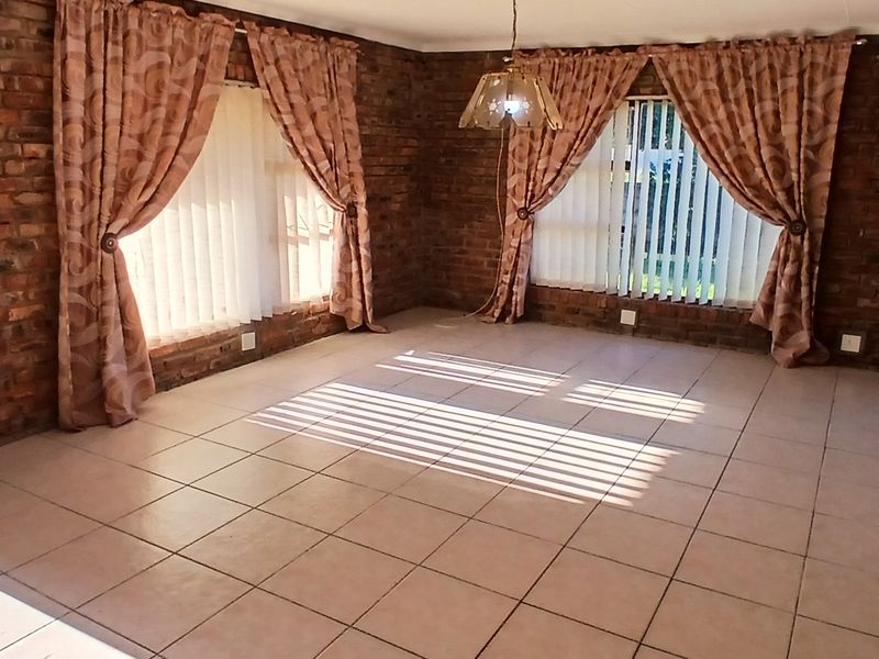 Spacious living area featuring exposed brick walls, ceramic tiled flooring, and large windows with vertical blinds, offering a bright and versatile living space.