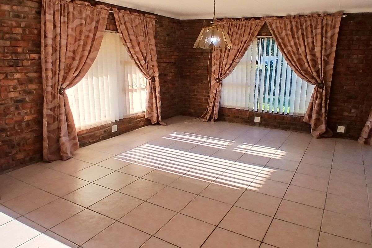 Spacious living area featuring exposed brick walls, ceramic tiled flooring, and large windows with vertical blinds, offering a bright and versatile living space.