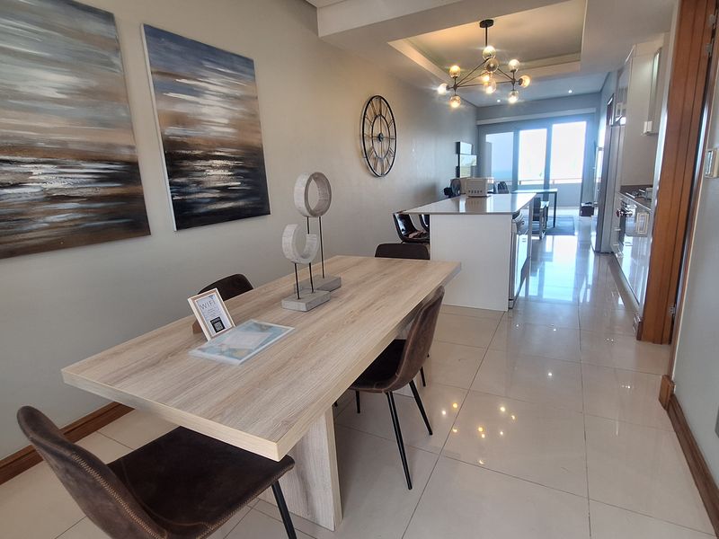Modern open-plan living and dining area featuring a central kitchen island, sleek tiled flooring, and recessed ceiling lighting, leading to a balcony.