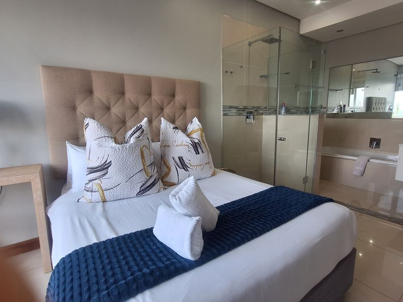 Modern bedroom featuring an open-plan en-suite bathroom with a glass-enclosed shower, built-in bathtub, and sleek tiled flooring.