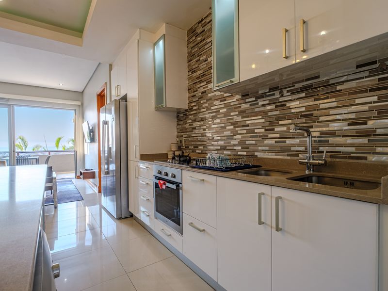 Modern kitchen featuring white cabinetry, stone countertops, a mosaic tile backsplash, and integrated stainless steel appliances, flowing into an open-plan living area.