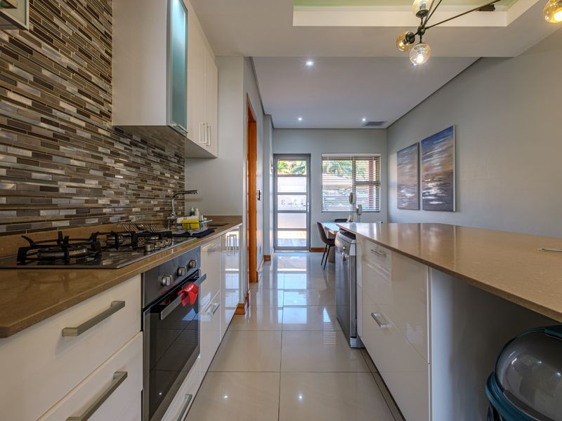 Modern open-plan kitchen featuring glossy cabinetry, stone countertops, a gas hob, and a breakfast bar, flowing into a bright dining area with tiled flooring.