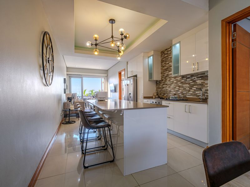 Modern open-plan kitchen featuring a central island, stone countertops, mosaic backsplash, and sleek white cabinetry, seamlessly flowing into the living area.