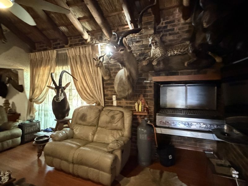 Rustic living room featuring exposed timber beams, a brick fireplace, and a built-in indoor braai area with timber flooring.
