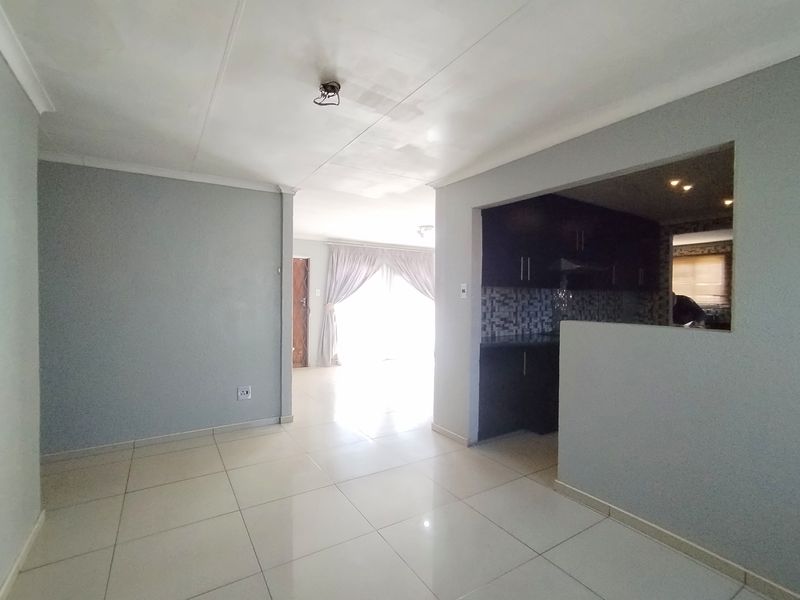 An open-plan living area featuring tiled flooring, a pass-through window to the kitchen, and neutral wall finishes, presenting a clean, vacant space.