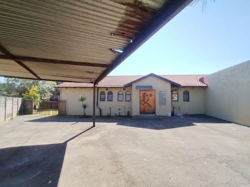 Front exterior view of the property featuring a paved driveway, a timber entrance door, and a corrugated iron carport structure.