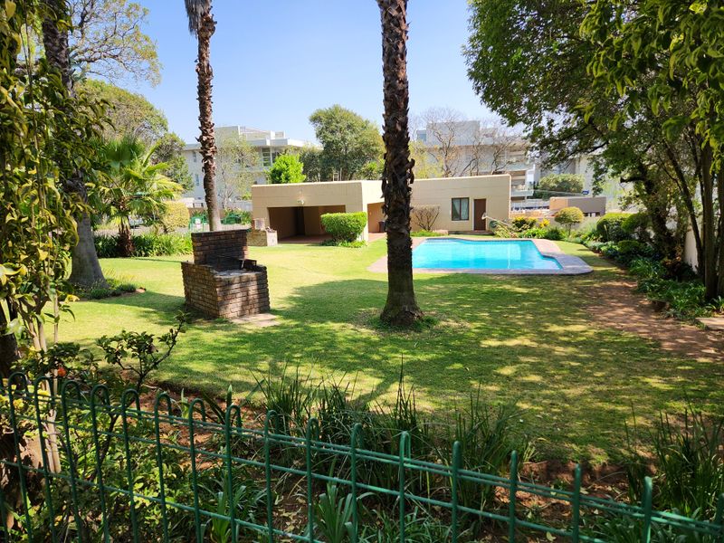 A well-maintained communal garden area featuring a swimming pool, brick built-in braai, and mature trees within a secure apartment complex setting.