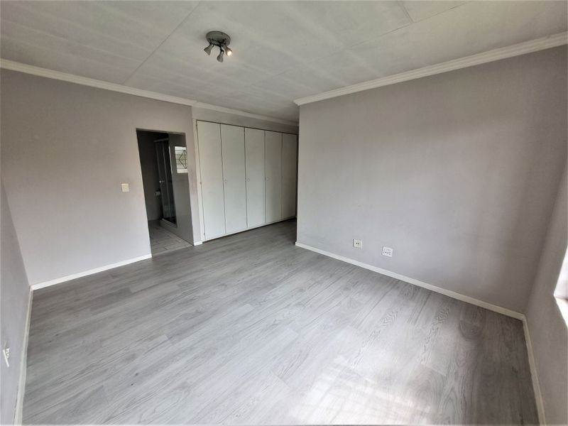 A spacious, empty bedroom featuring light-toned laminate flooring, built-in cupboards, and neutral wall finishes, with an adjoining en-suite bathroom area.