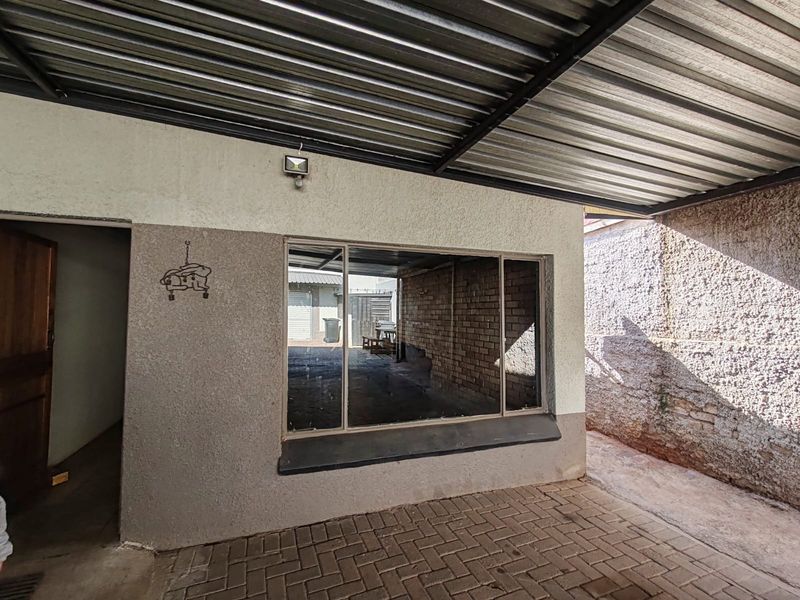 A covered outdoor patio area featuring paved flooring, a corrugated metal roof, and a large window looking into the property.