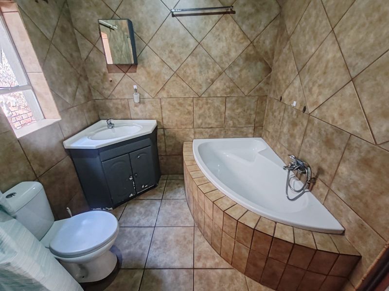 A functional bathroom featuring a corner bathtub, vanity cabinet with basin, and toilet, finished with full-height wall tiling and floor tiles.