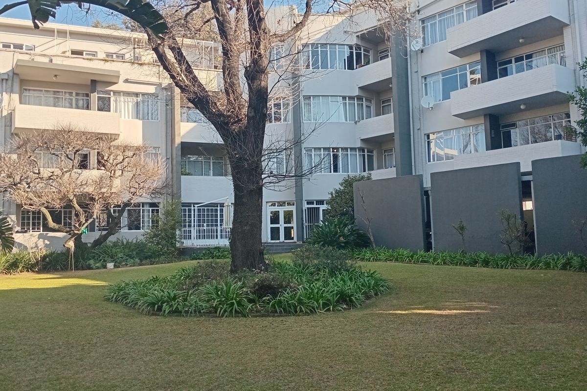 A view of the rear facade of the apartment building featuring a well-maintained communal garden with a large mature tree and private balconies.
