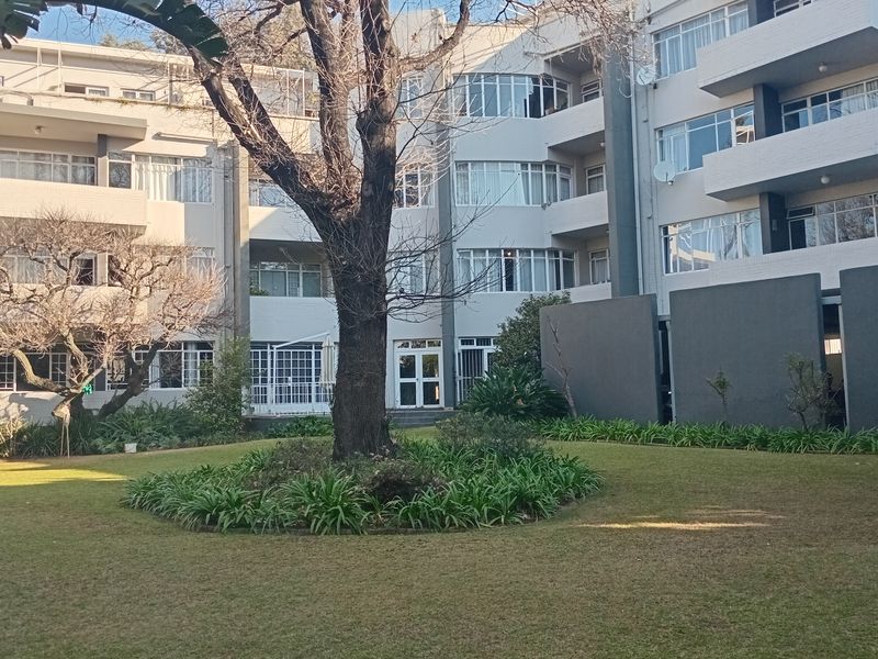 A view of the rear facade of the apartment building featuring a well-maintained communal garden with a large mature tree and private balconies.