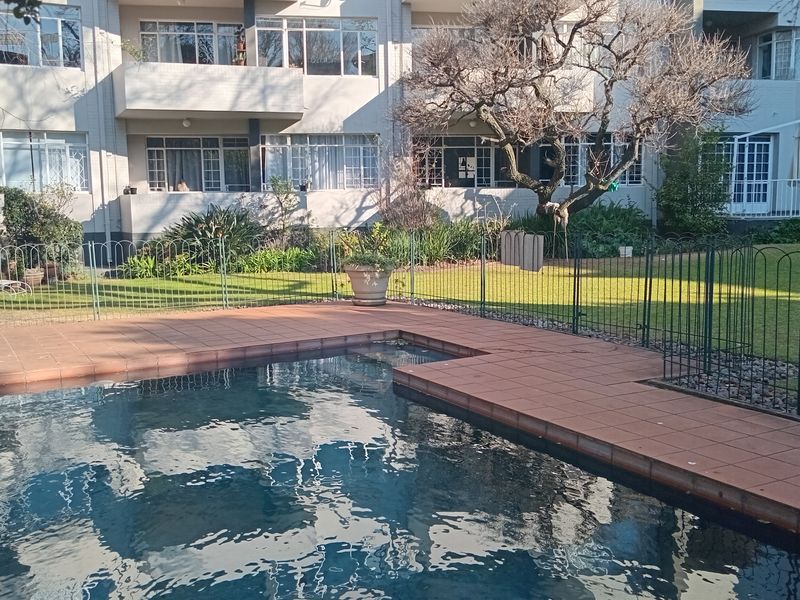A communal swimming pool area featuring a tiled surround and metal fencing, set within a well-maintained garden space of a flat complex.