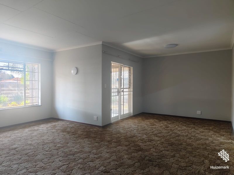 A spacious, empty living room featuring neutral walls, carpeted flooring, and a security gate leading to the exterior.
