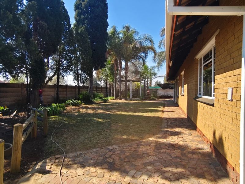 A well-maintained rear garden featuring a brick-paved walkway, established trees, and a face brick exterior wall with guttering.