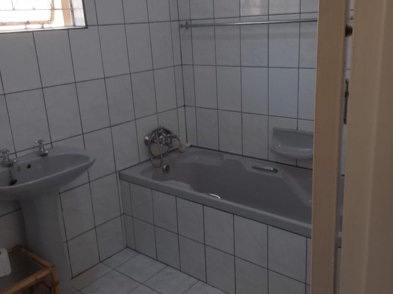A functional bathroom featuring a pedestal basin, built-in bathtub, and full-height wall tiling with a window for natural light.