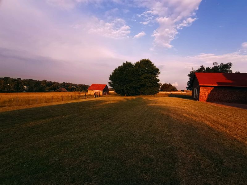 A wide view of the property grounds featuring a large lawn, brick outbuildings with metal roofs, and surrounding open fields at sunset.