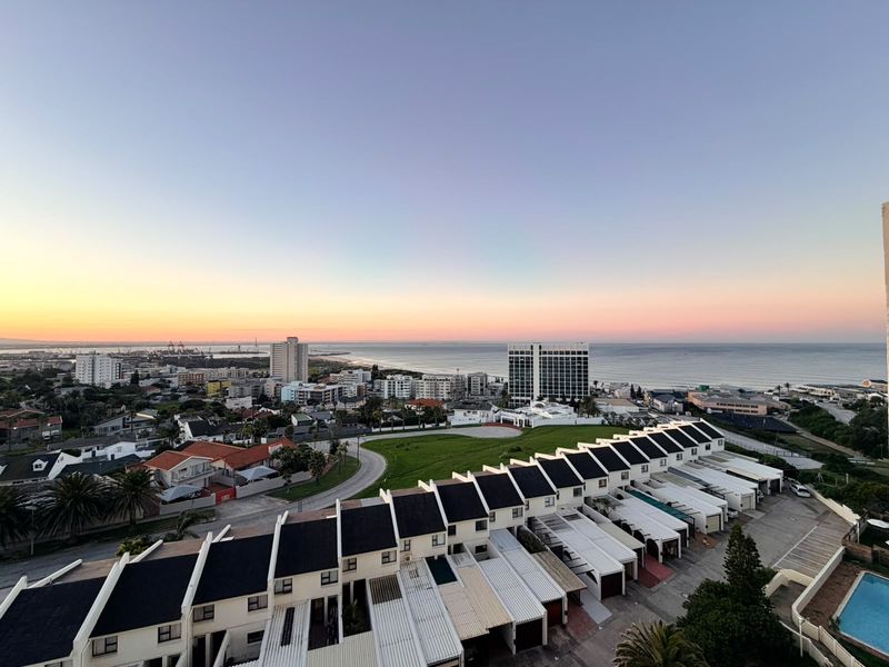 Elevated panoramic view overlooking the coastal neighbourhood, residential townhouses, and the ocean at sunset.