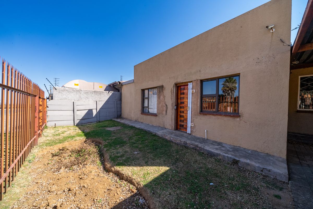 Rear exterior view of the property featuring a concrete walkway, security fencing, and a small garden area with visible plaster wear.