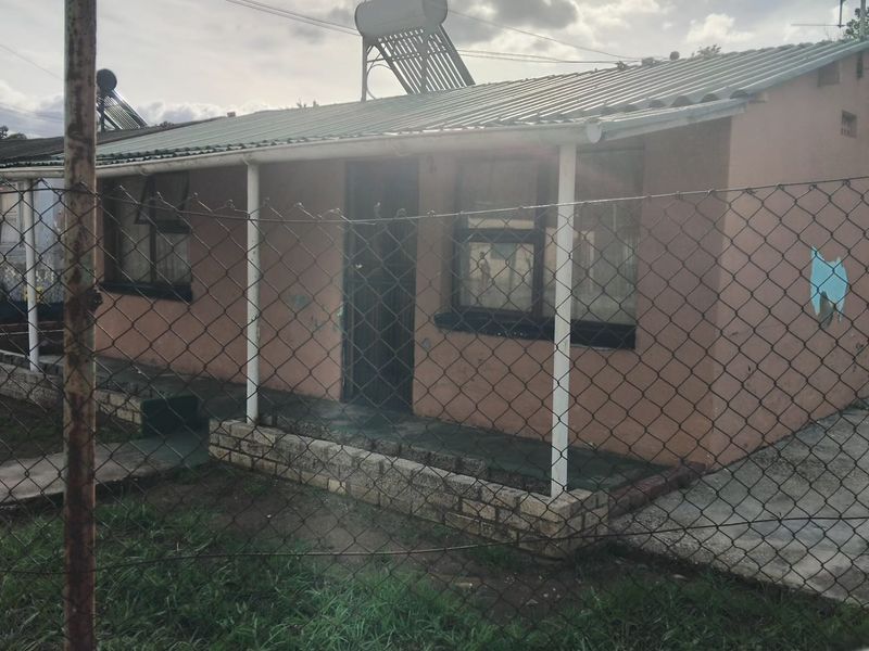 Front view of a single-story house featuring a corrugated metal roof, solar water heater, and a covered porch area enclosed by a chain link fence.