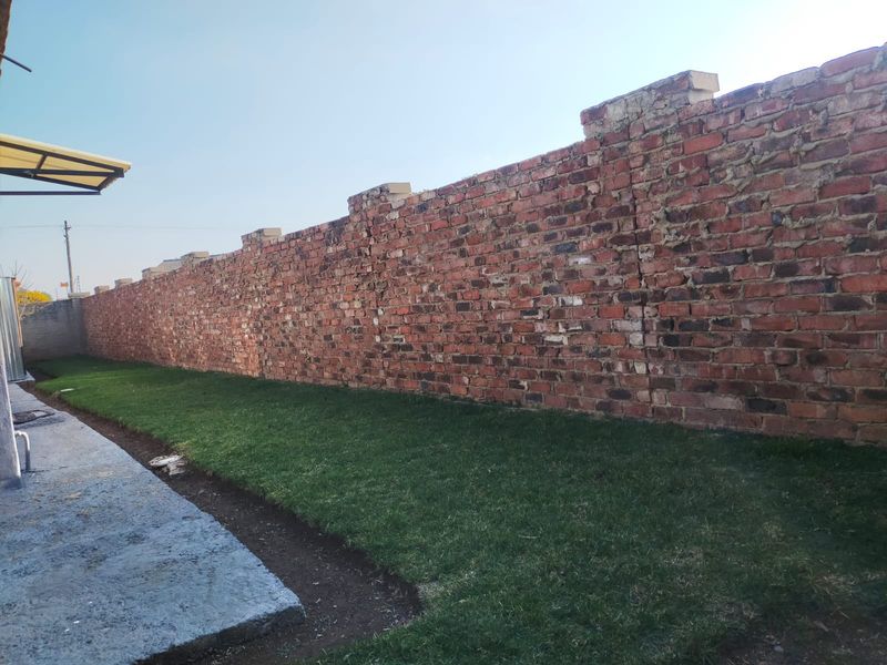 A rear garden area featuring a long, weathered brick boundary wall, a small lawn, and a concrete pathway.