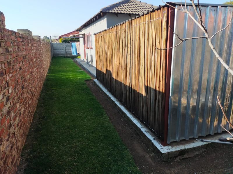 A side yard view featuring a brick boundary wall, a corrugated metal structure with reed screening, and a small lawn area.