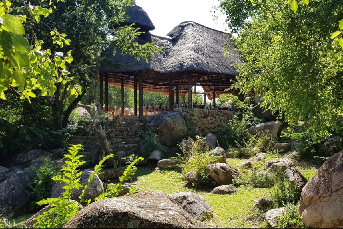 A scenic view of a lodge structure featuring a traditional thatched roof, timber pillars, and stone masonry, set within a natural rock garden landscape.