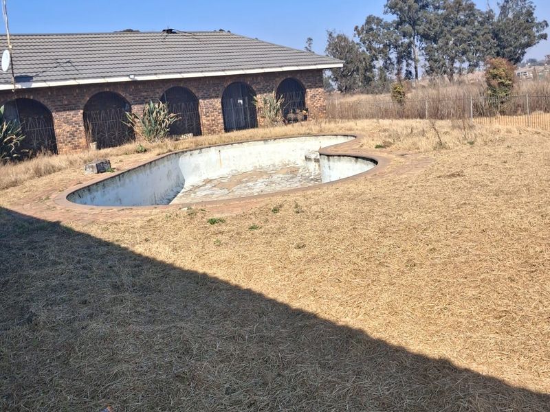Exterior view of a backyard featuring an empty swimming pool and a brick house with arched architectural details, set in a dry, unmaintained garden.