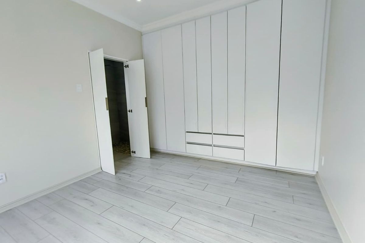 A bright, empty bedroom featuring modern white built-in cupboards, light-toned laminate flooring, and recessed ceiling lighting.