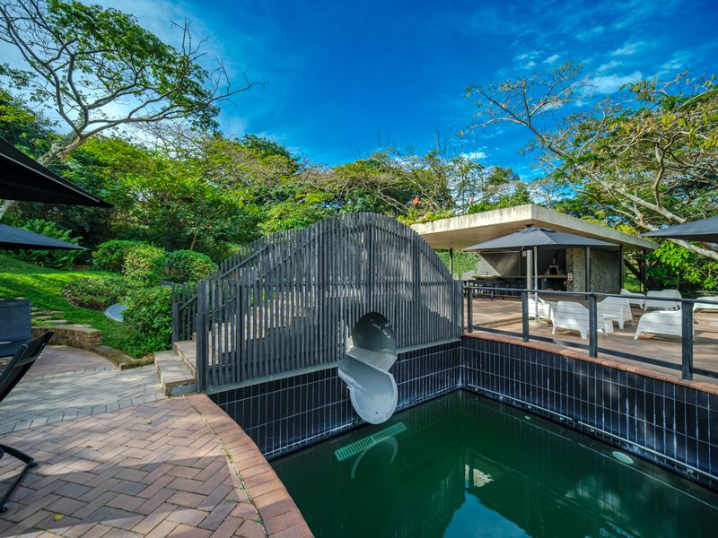 Outdoor pool area featuring a built-in slide, timber decking, and a covered patio with an outdoor kitchen setup, surrounded by lush garden landscaping.