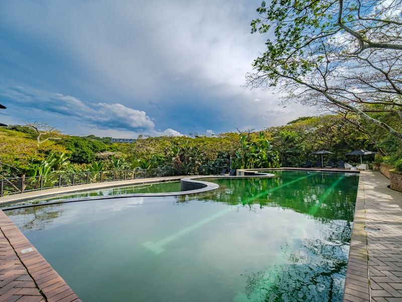 A large outdoor swimming pool featuring brick paved surrounds and metal safety railings, set within a lush, landscaped garden environment.