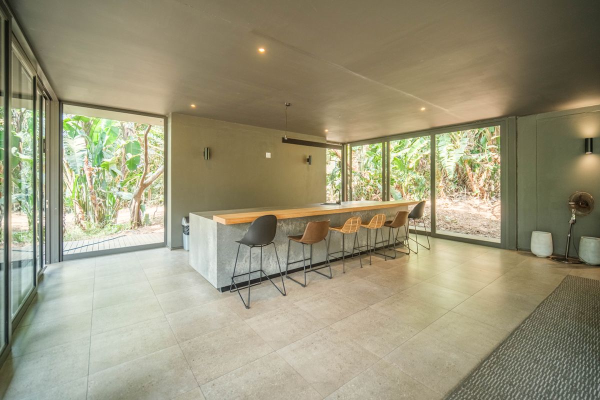 Modern open-plan kitchen featuring a large concrete island with a wooden breakfast bar, set against expansive glass walls overlooking lush greenery.